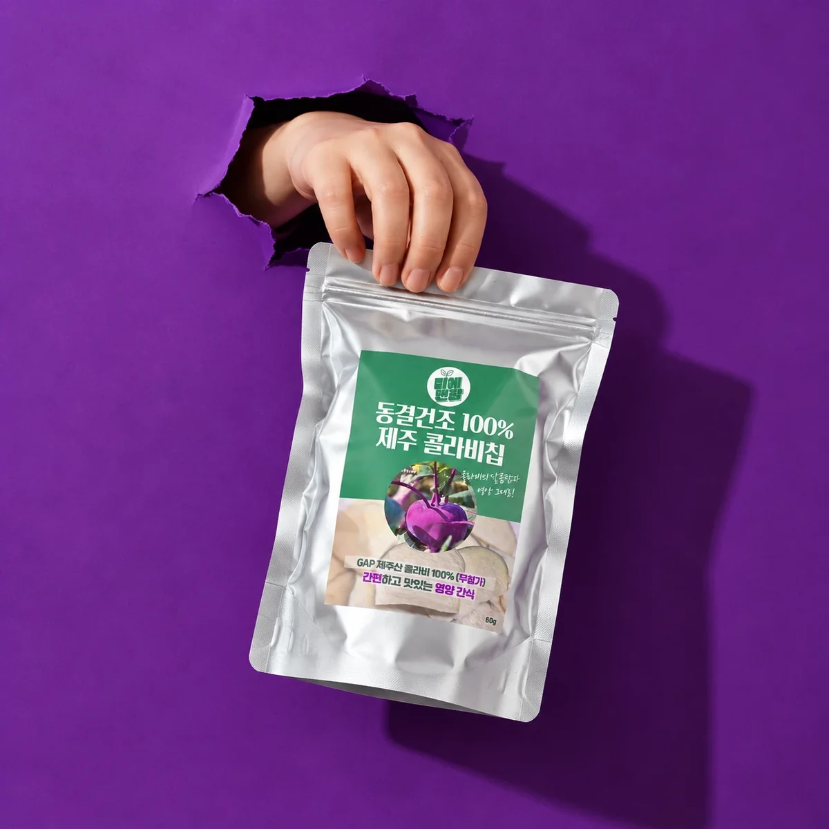 A hand bursting through purple paper, holding a pack of Jeju kohlrabi chips